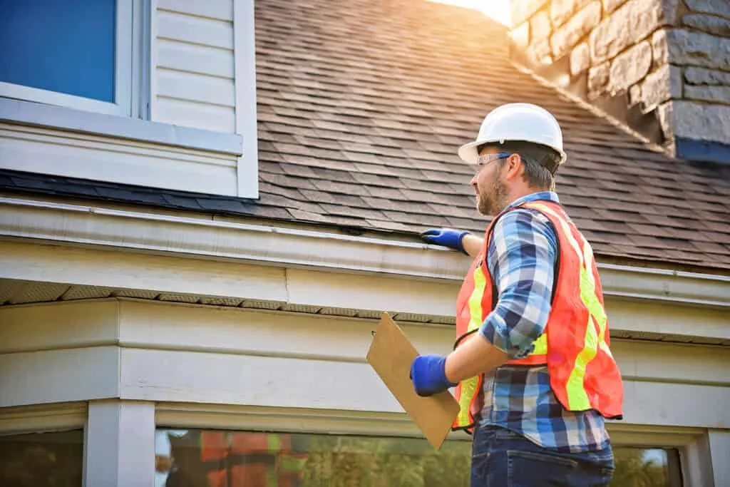 A worker in a hard hat and safety vest writing on a clipboard beside a house